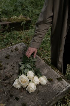 A solemn moment captured as a hand places white roses on a moss-covered tombstone, symbolizing loss and bereavement.